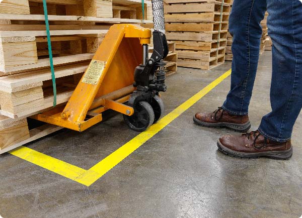 Gul ToughStripe-gulvtape er påsat gulvet på et lager. Tapens position viser, hvor en medarbejder med en palleløfter skal placere en stak materialer.